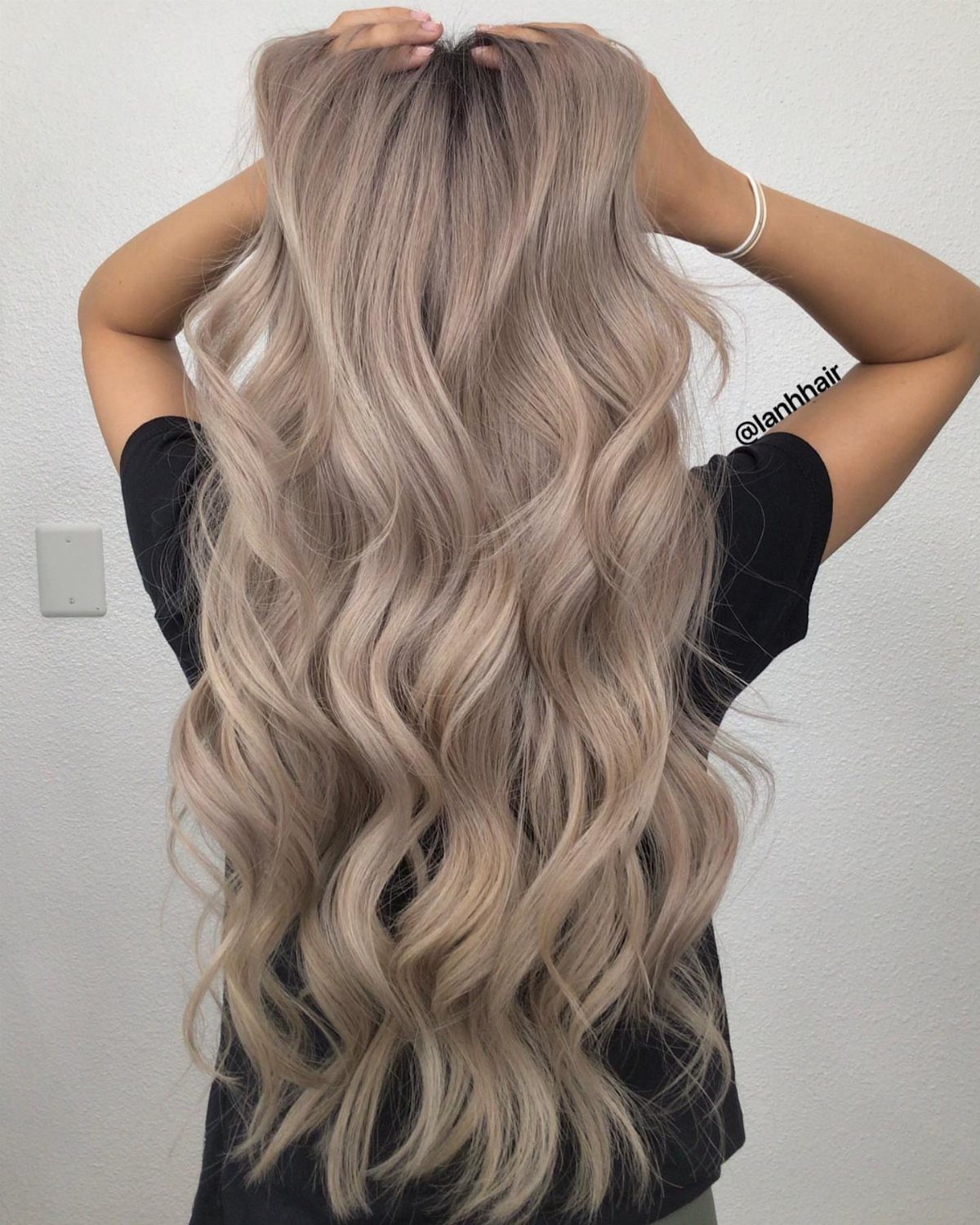 Long ash blonde waves cascade down the back of a person in a black shirt