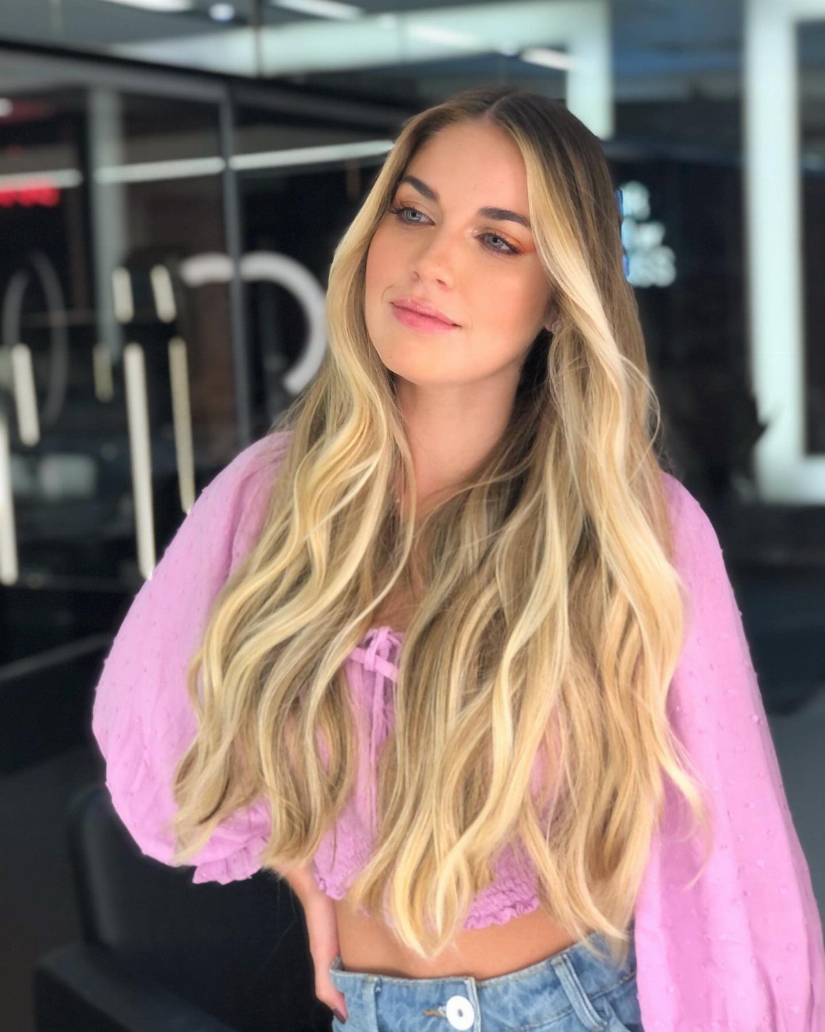 Woman with long, wavy blonde hair and darker roots wearing a pink top