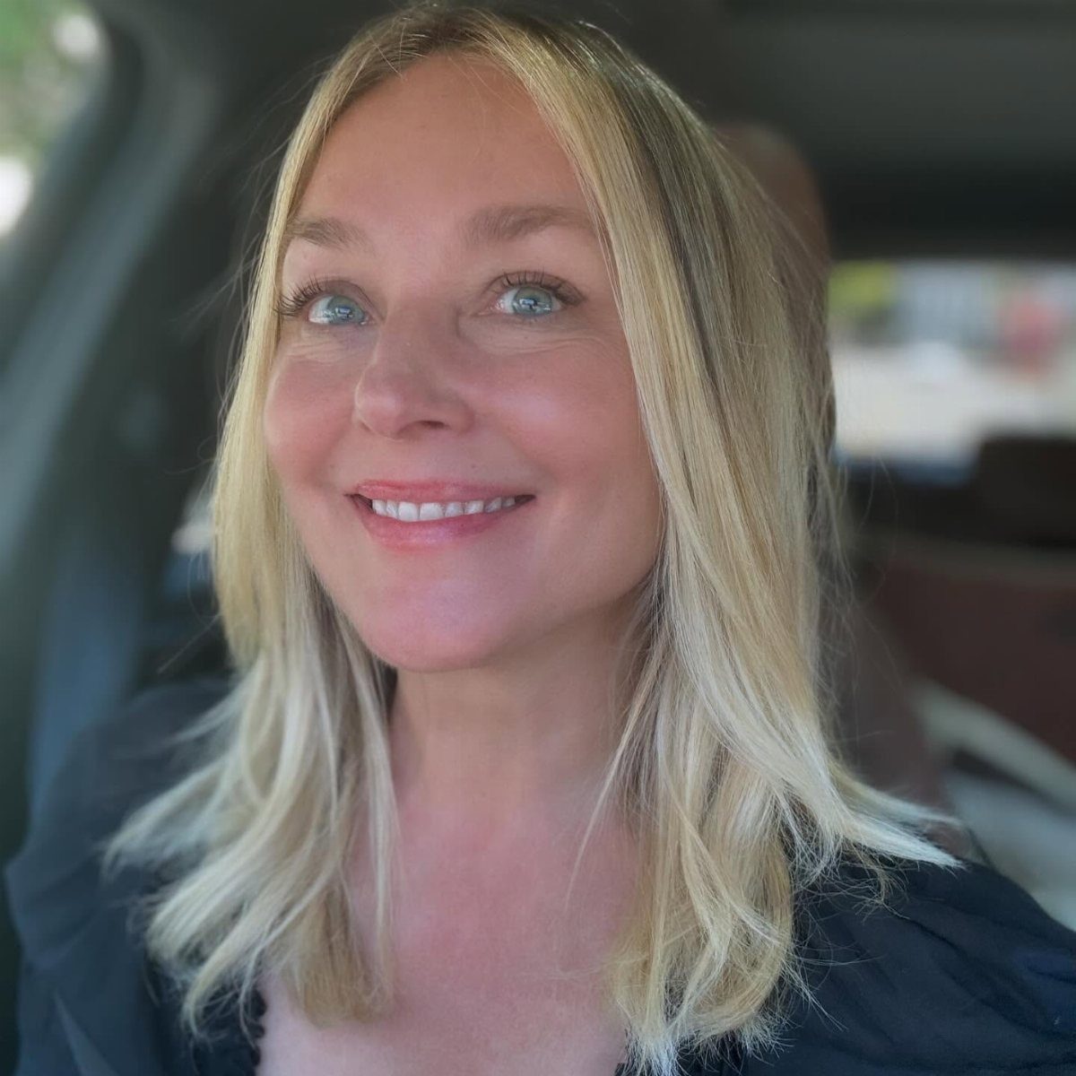 Woman with long, dark-rooted ash-blonde hair smiling inside a car