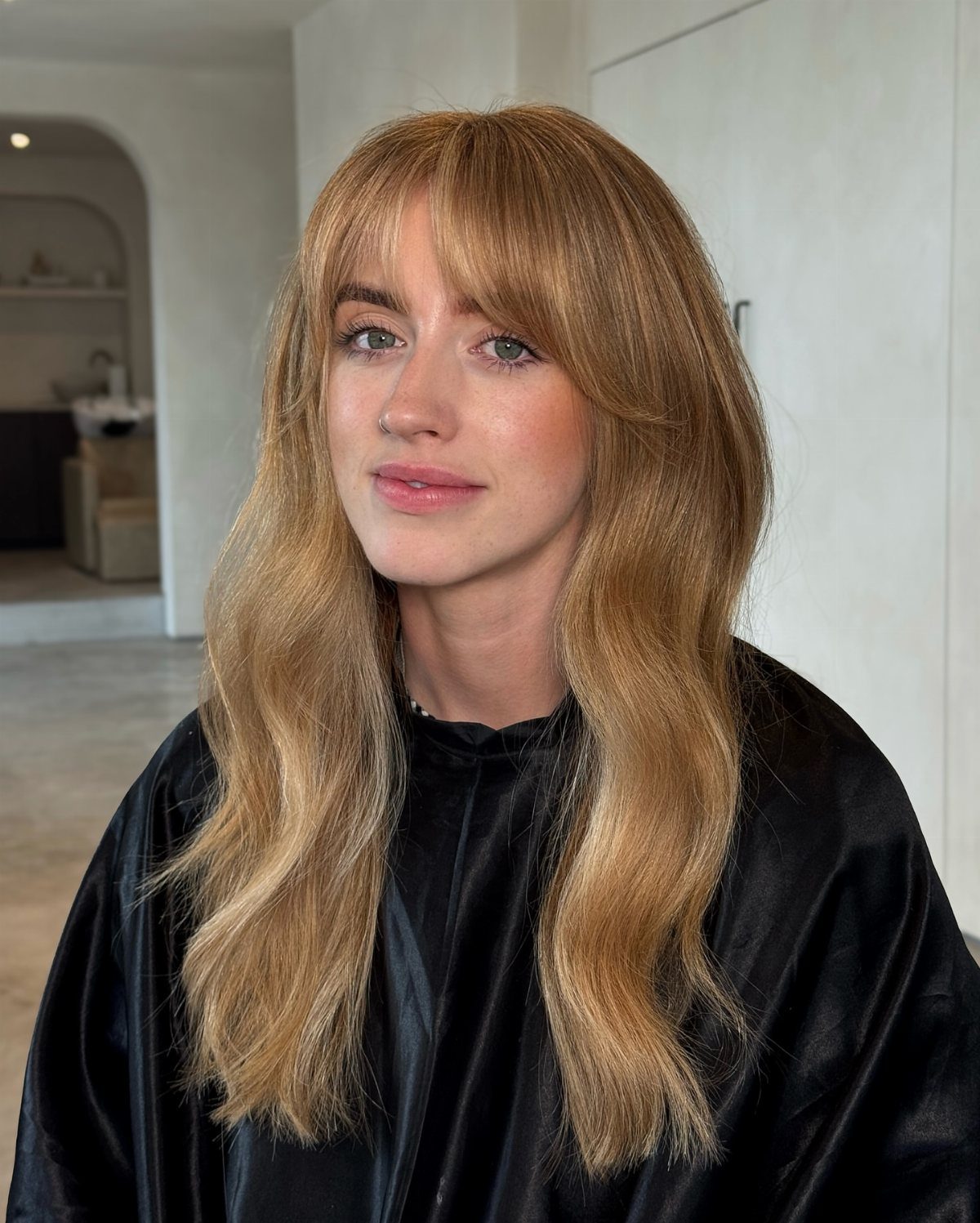 Woman with long, wavy dark blonde hair and bangs wearing a black salon cape